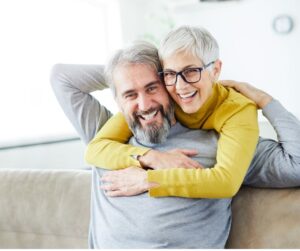 This is an image of a older couple. The man is seated on a couch and the woman is hugging him from behind. 