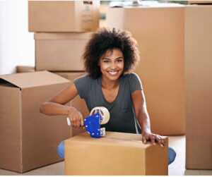 This is an image of a woman sitting on the floor, surrounded by boxes, holding a tape gun to tape up a box.